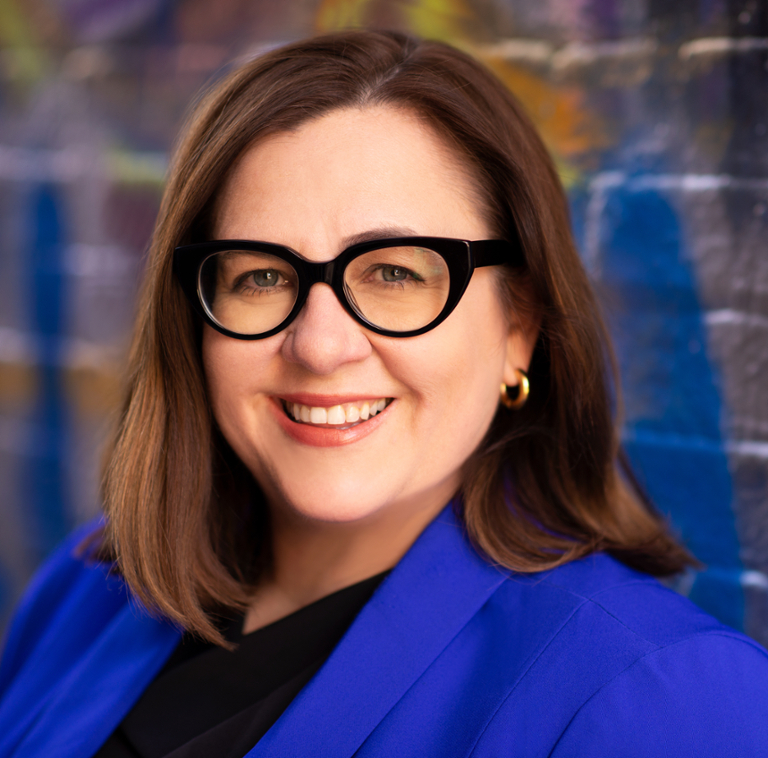 Woman in a blue blazer with thick-rimmed glasses standing in front of a colorful wall.